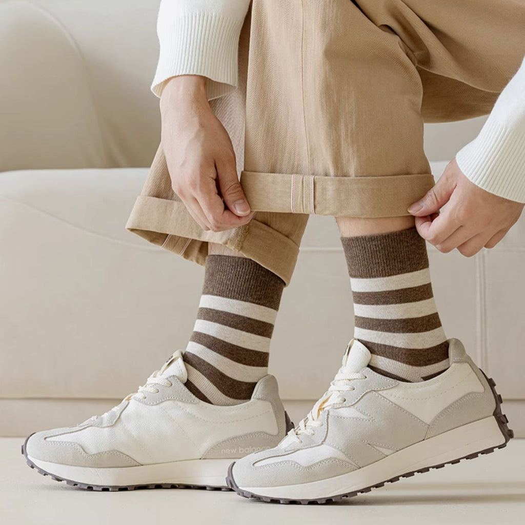 A person seated on a light sofa wearing beige pants, a white sweater, brown and beige striped socks, and white sneakers.