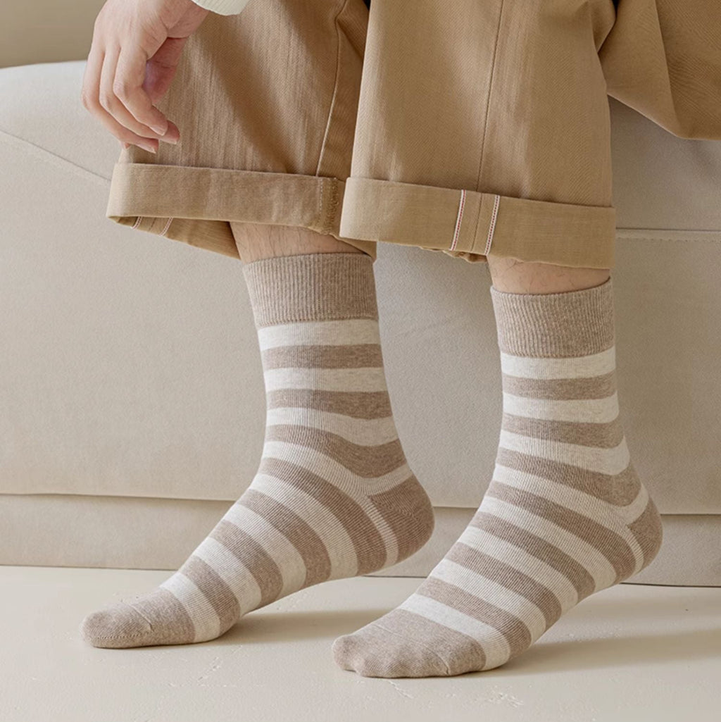 A person seated indoors wearing beige and off-white striped crew socks with tan pants and a cream sweater. The photo is cropped from the waist down, with the socks clearly visible on both feet.