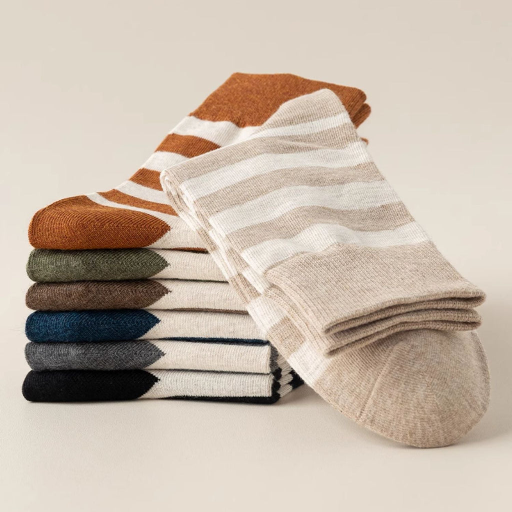 A stack of folded knit socks in earthy tones, with one beige sock featuring white horizontal stripes draped over the front. The visible colors in the stack include rust orange, olive green, navy blue, charcoal gray, and black.