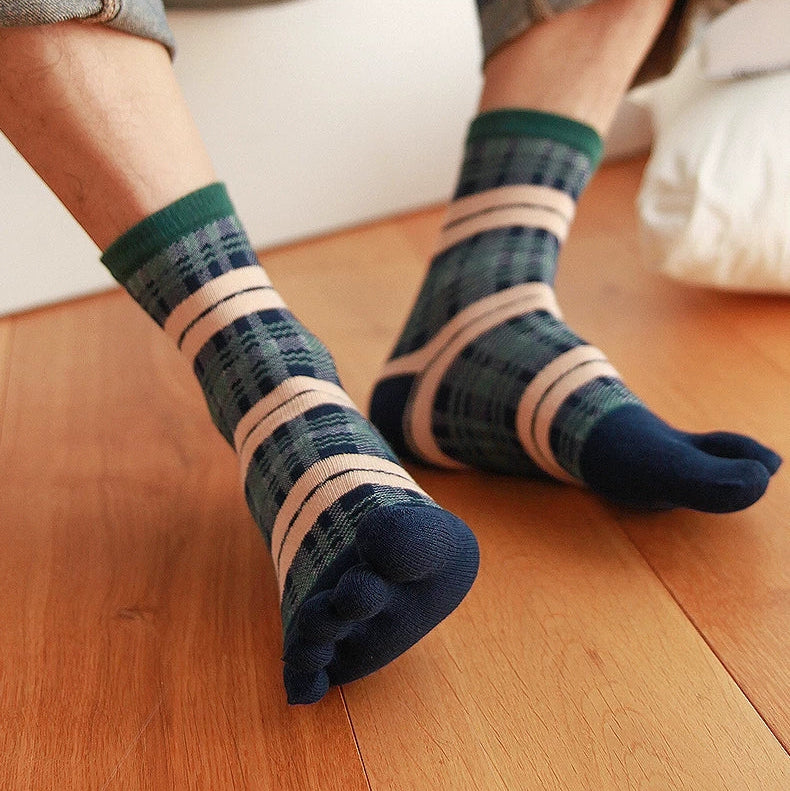 A person sitting on a wooden floor wearing striped socks with a plaid pattern in navy, green, and beige, paired with rolled-up denim jeans.