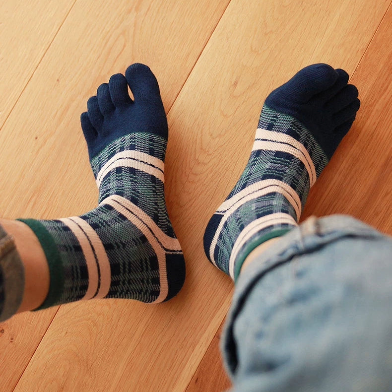 A pair of striped and plaid five-toe socks worn on feet, shown from above on a light wood plank floor with light blue jeans visible at the bottom right.