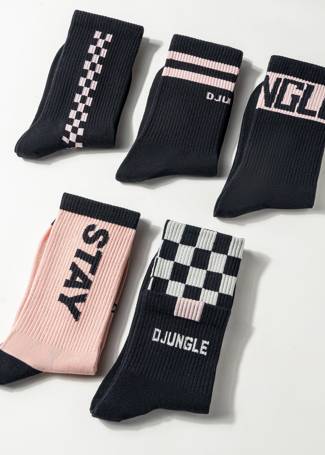 Five pairs of crew socks arranged on a light gray background. The socks are primarily black with pink and white accents, featuring checkerboard patterns, stripes, and text including 'STAY', 'DJJUNGLE', and 'NGL'.