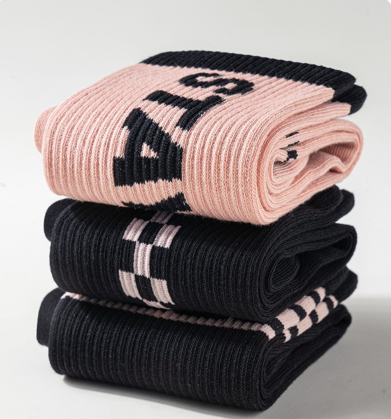 Three folded ribbed knit socks stacked on a light gray background, with Chinese text at the bottom left. The top sock is light pink with large black lettering, the middle sock is black with light pink stripes, and the bottom sock is black with a black-and-pink check pattern.
