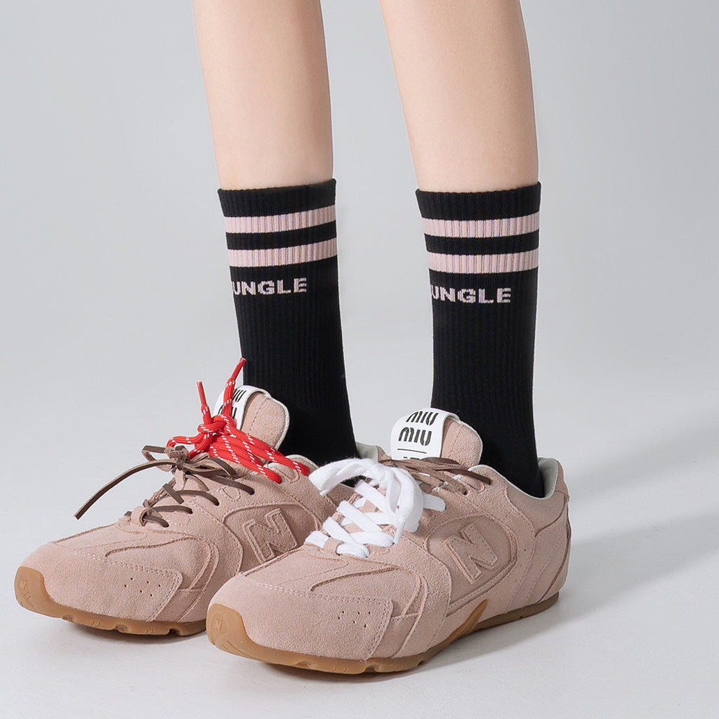 A person wearing beige suede sneakers with white and red laces, paired with black crew socks featuring pink stripes and the word 'MINGLE' in white, shown against a light gray studio background.