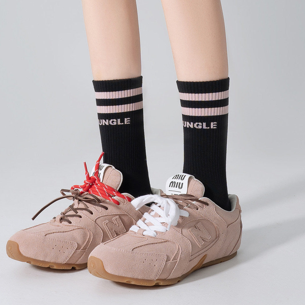 A person wearing beige suede sneakers with white and red laces, paired with black crew socks featuring pink stripes and the word 'MINGLE' in white, shown against a light gray studio background.