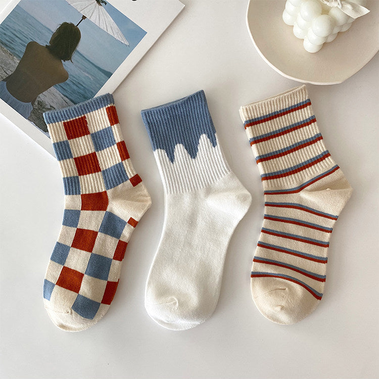 Three pairs of ankle socks displayed on a light surface: one pair with a red, blue, and cream checkerboard pattern; one pair with a blue ribbed cuff and cream body; and one pair with cream, red, and blue horizontal stripes.