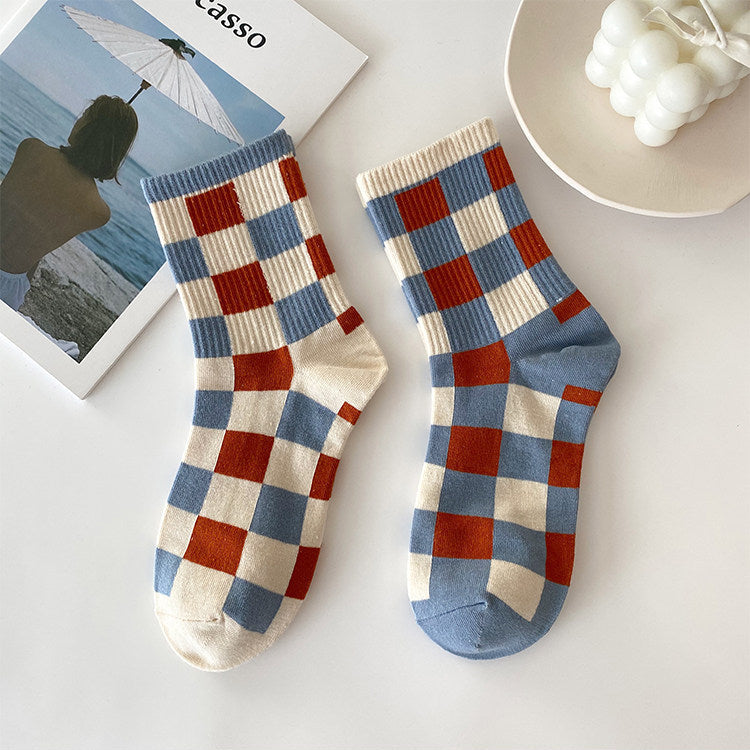 A pair of ankle socks with a blue, red, and cream checkerboard pattern, styled on a white surface with a magazine and a bowl of white candles.