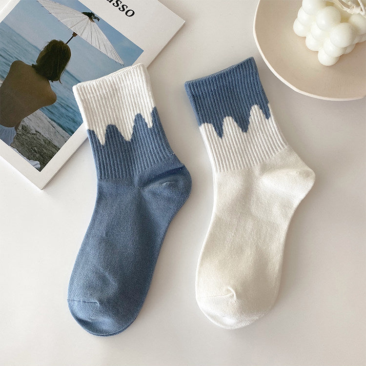 Two ankle socks displayed on a light surface, one in dusty blue and one in off-white, both featuring a wavy dip-dye style cuff. A magazine and a bowl of white grapes are styled props in the background.