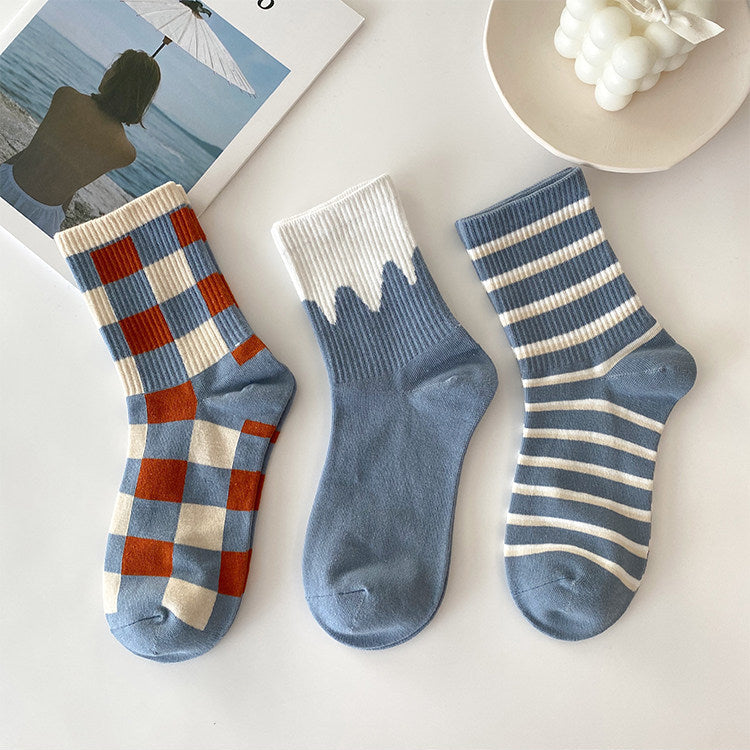 Three pairs of ankle socks arranged on a light surface: one blue, red, and cream check pattern; one dusty blue with a cream wavy cuff; one dusty blue with cream horizontal stripes. A magazine page and a small dish are partially visible as styled props.