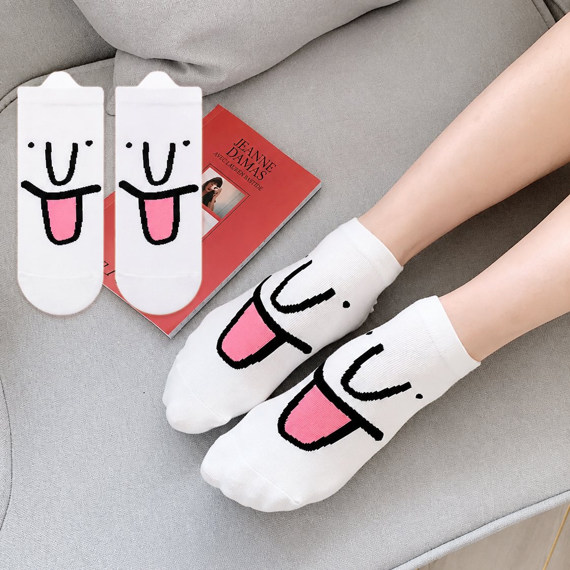 White socks with pink tongue design worn by a person on a gray couch, with packaging in the background.