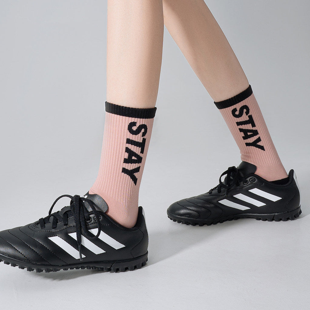 A person's legs wearing light pink crew socks with black trim and the word 'STAY' printed vertically, paired with black soccer cleats with white stripes, against a light gray studio background.